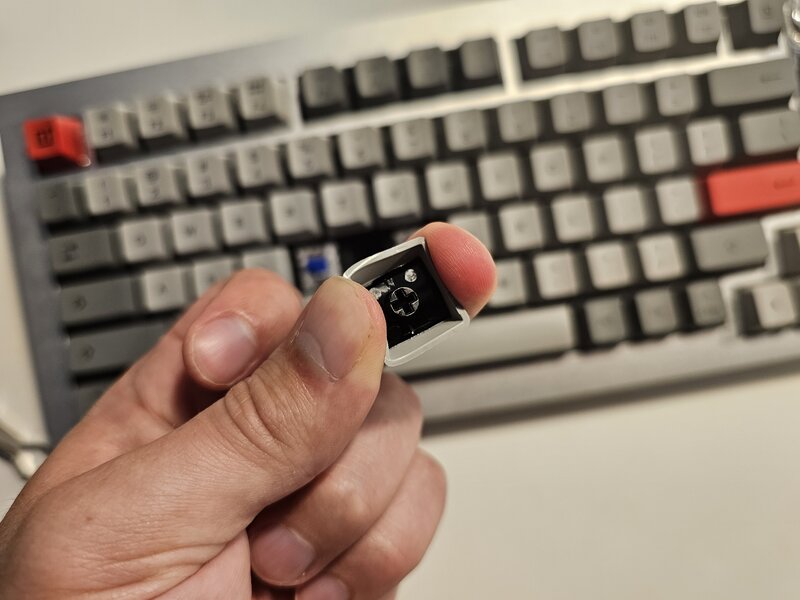 OnePlus Keyboard 81 Pron näppäimistö taustalla, edessä sormien välissä näppäinhattu