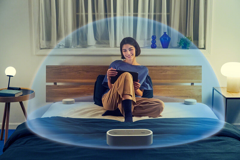 Nainen istuu sängyllä kuuntelemassa 360 asteen ääntä, jonka Sony HT AX7 kannettava kaiutinjärjestelmä luo käyttäjän ympärille