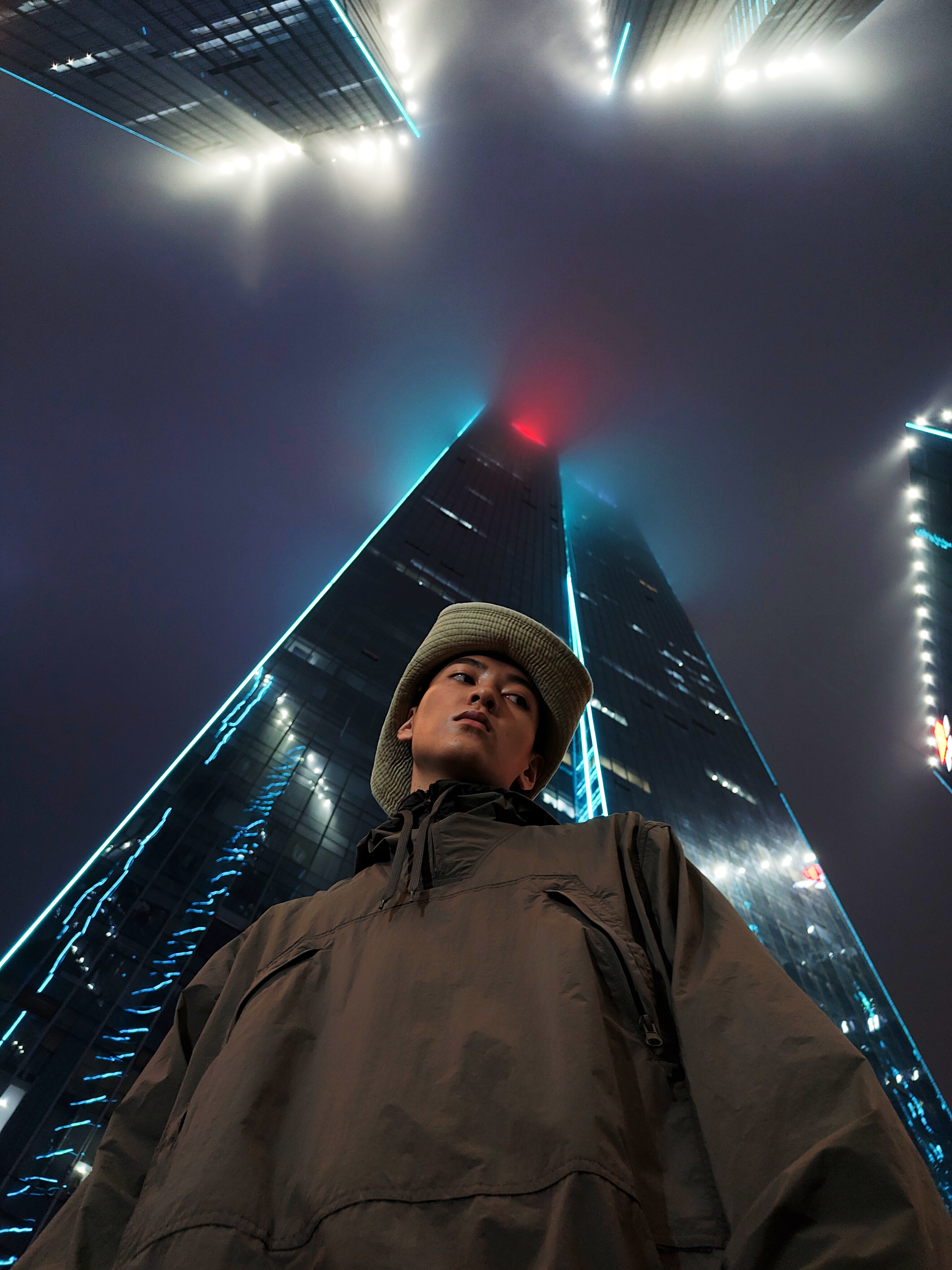 alaviistosta otettua kuva ruskeatakkisesta miehestä jonka taustalla on korkeita rakennuksia sumuisessa ja pimeässä illassa